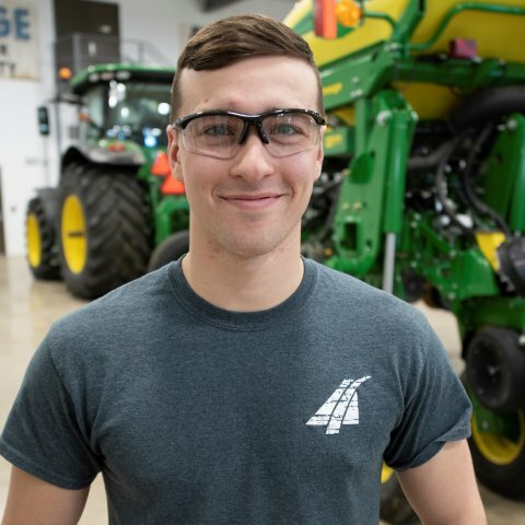A student wearing safety glasses stands smiling in front of a tractor