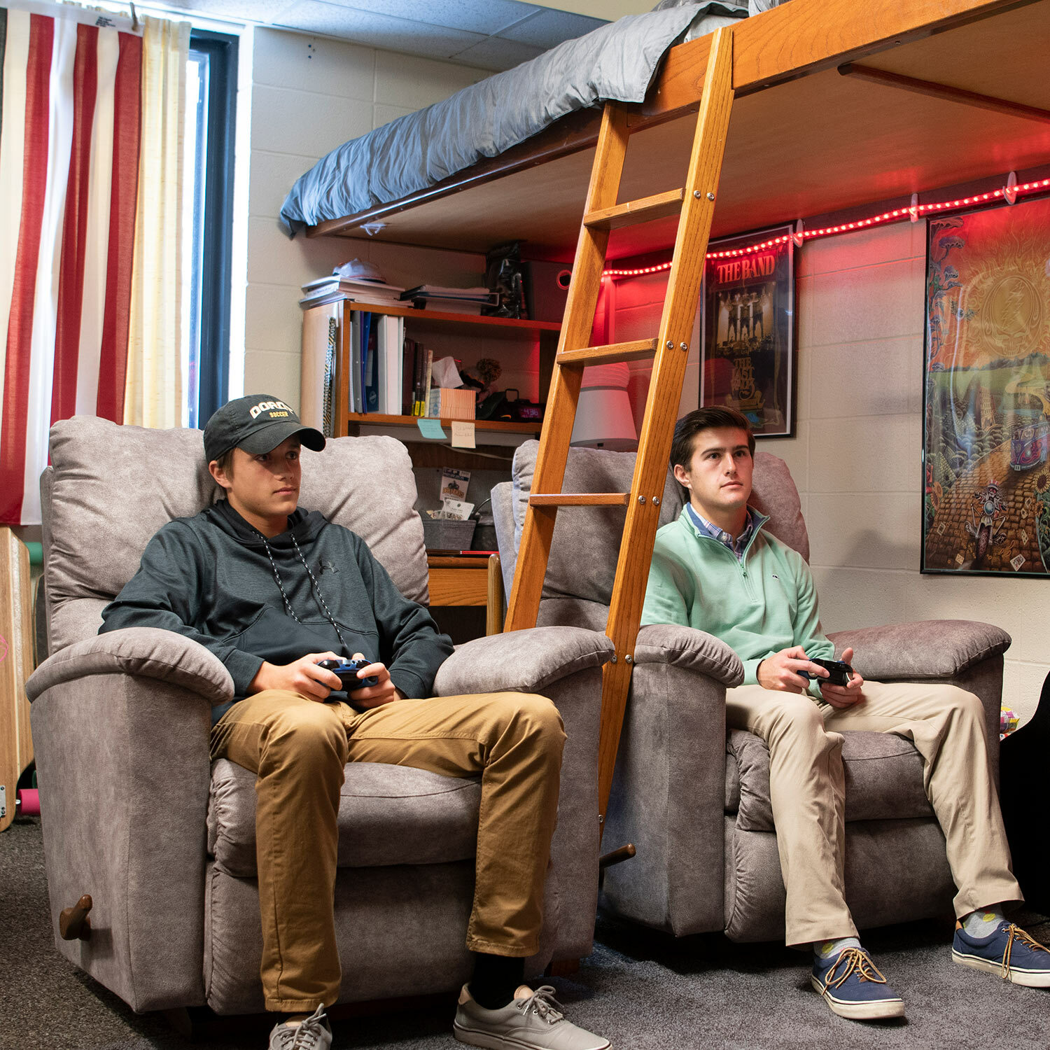 Two young men sit in recliners in dorm room with video game controllers in hand