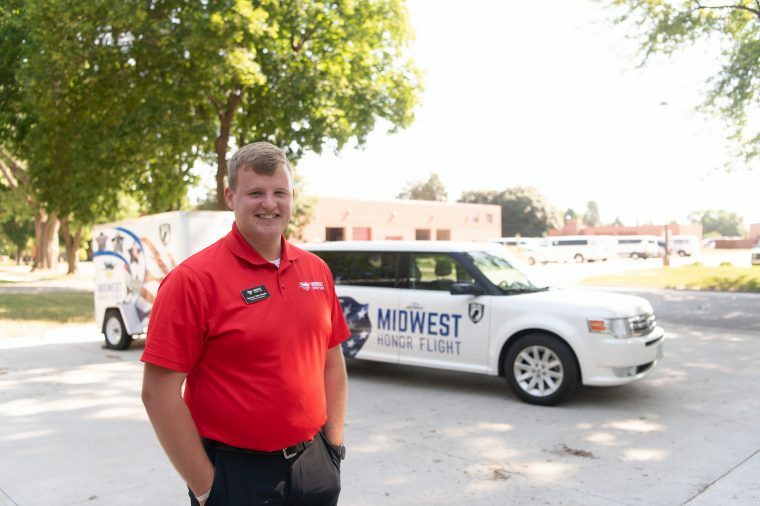 A Midwest Honor Flight organizer stands in front of a vehicle and trailer bearing the Midwest Honor Flight logo