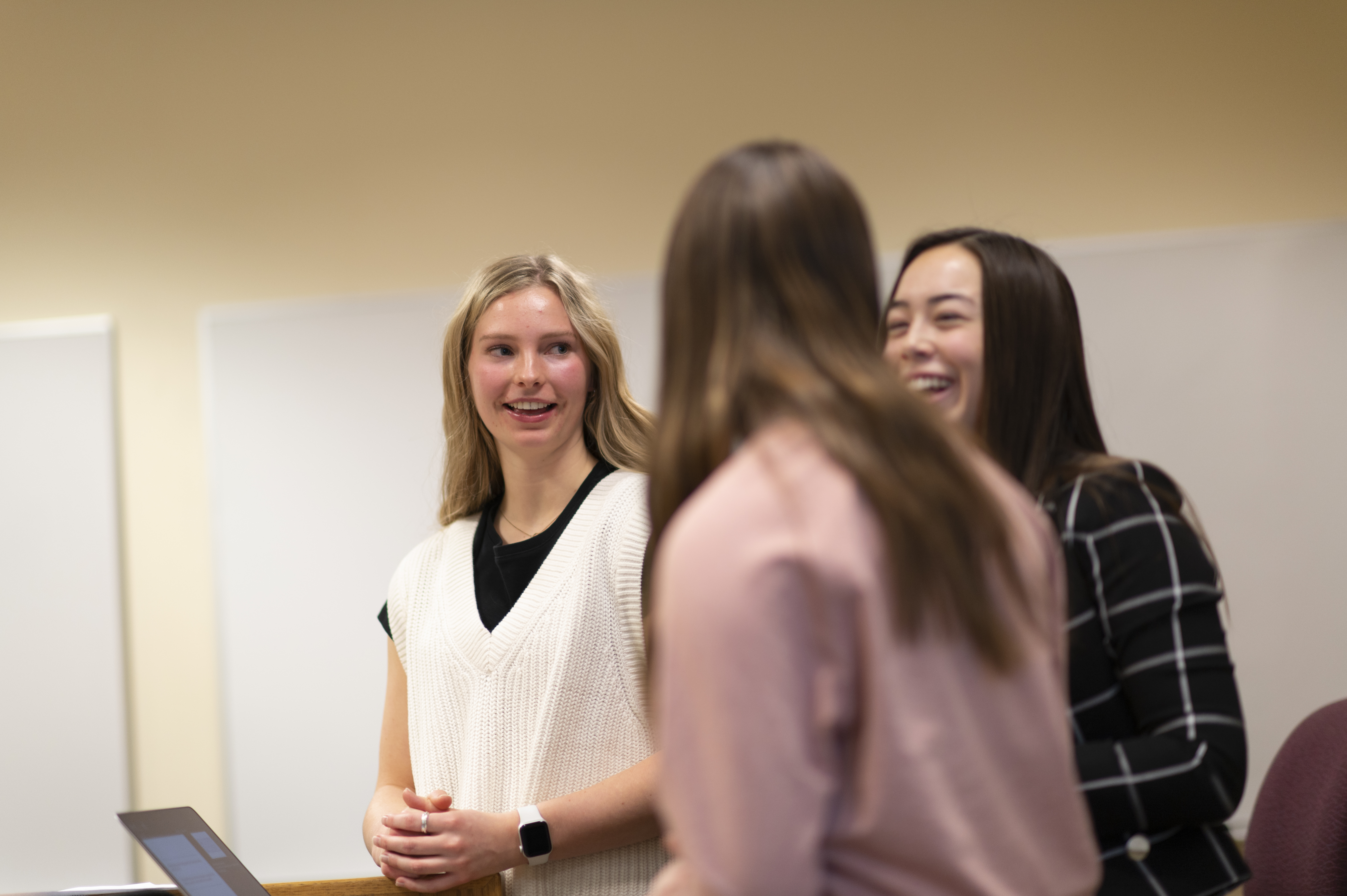 Three female students smile at each other while presenting to the class