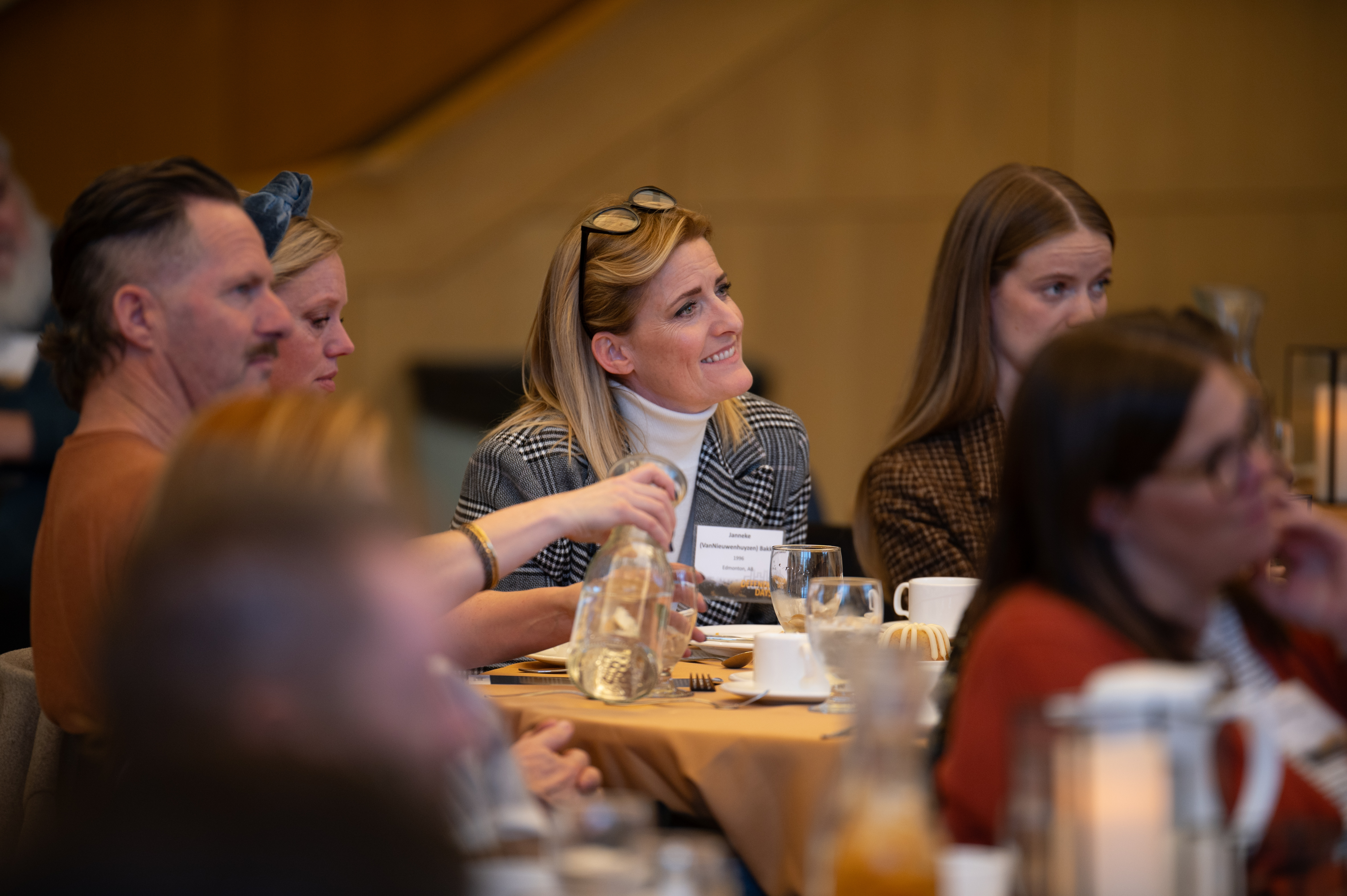 A diverse group of people sitting at a table, attentively listening during an indoor event. The setting appears formal, with table decorations and glasses. The focus is on a smiling woman with glasses on her head, surrounded by others.