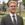 Photo of young smiling blond man in suit and tie with campus in the background