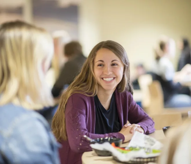 A women in a purple jacket smiles at her friend over lunch