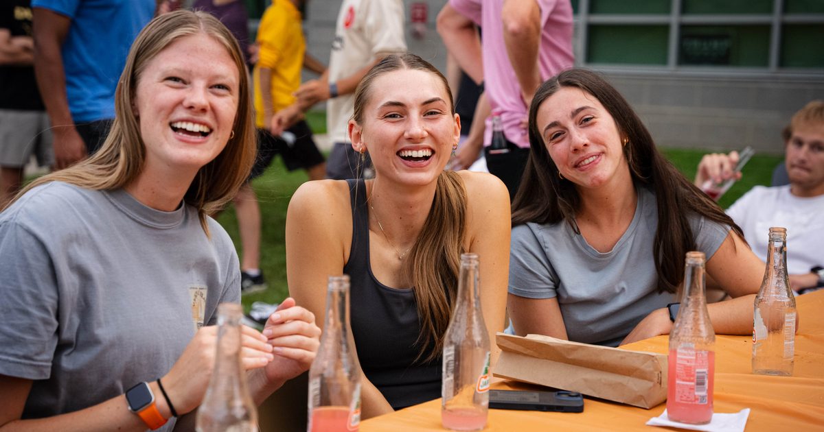 Three young women sit at a table outdoors, smiling and laughing. Empty and half-full glass soda bottles are on the orange tablecloth in front of them. Other people are visible in the background.