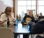 Two female students laugh at a round table while enjoying food in a large, window-filled space