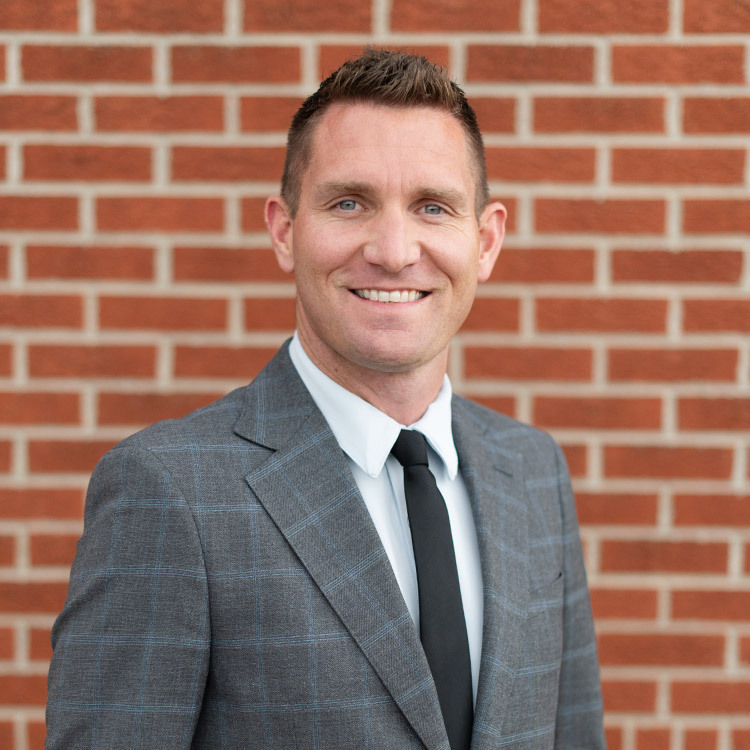 Man dressed in suit and tie standing and smiling in front of brick wall outdoors