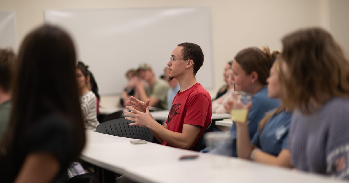 A man in a red shirt speaks while seated at a classroom table among other students, who are listening attentively. A whiteboard is visible in the background.