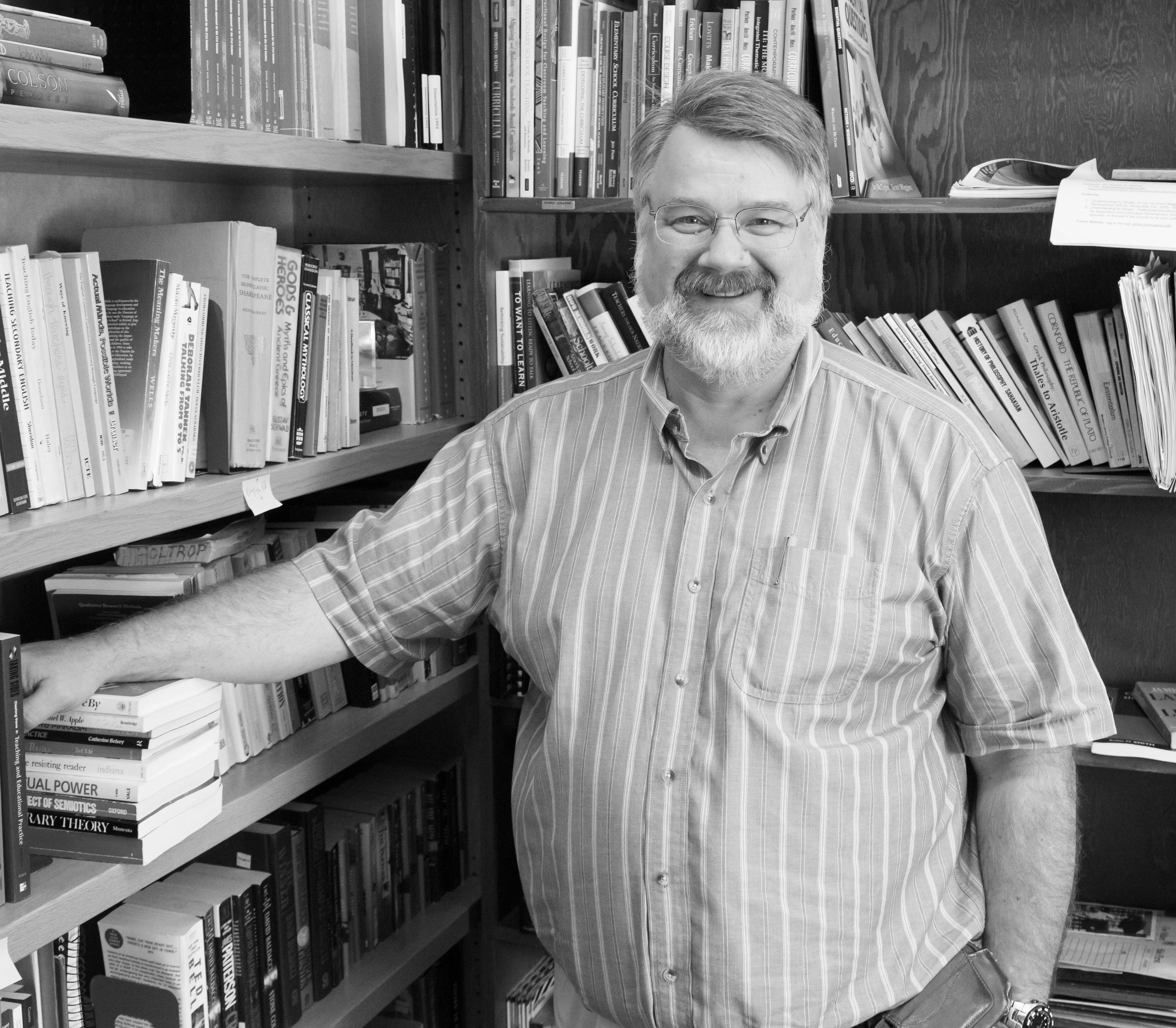 Black and white photo of bearded man standing in corner of room amidst bookshelves