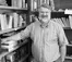 Black and white photo of bearded man standing in corner of room amidst bookshelves