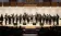 A group of musicians dressed in black performs on stage with music stands and microphones, while an audience watches from the seats in a concert hall.