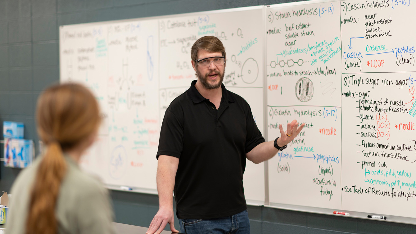 A Professor in safety glasses in front of a white board giving a lecture