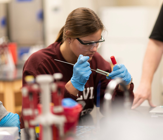 A Dordt student with safety glasses and gloves on looking into a test tube