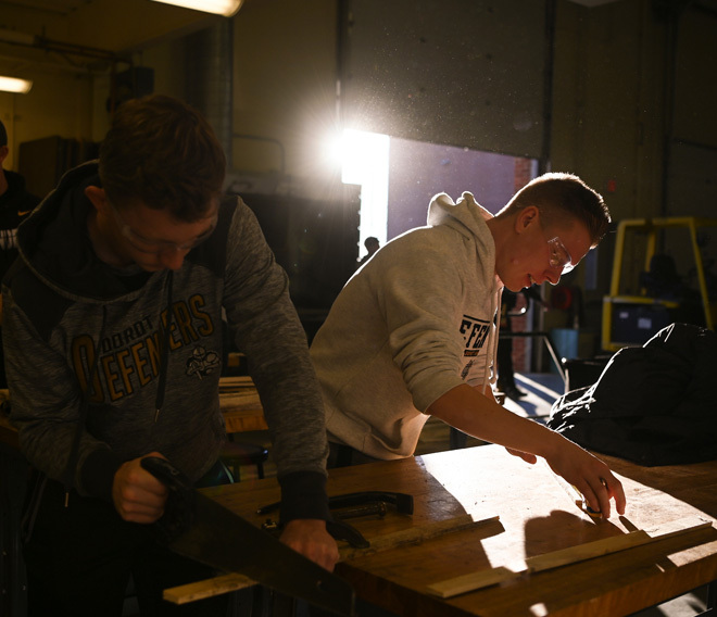 Two Dordt Students standing up with safety glasses on as one is measuring a piece of wood the other is about to saw it