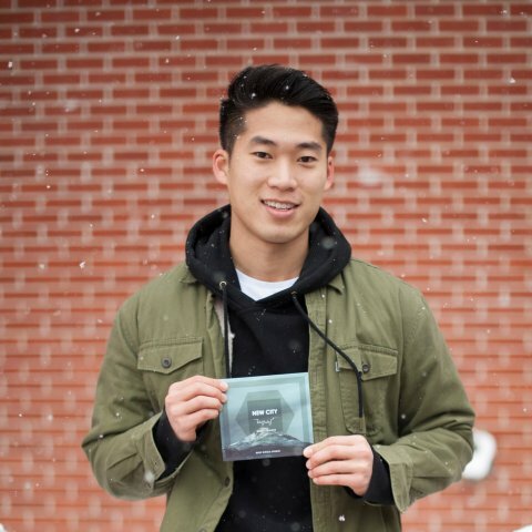 A man in a black hoodie and green jacket stands in the snow holding up a picture
