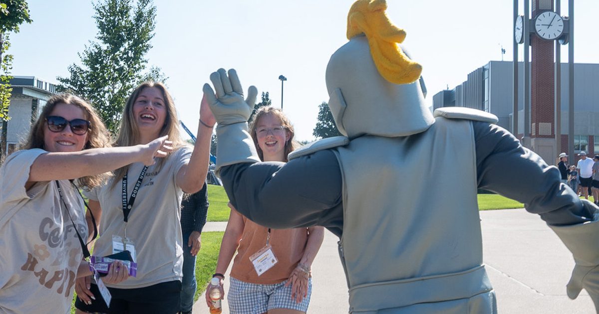 A person in a knight mascot costume high-fives three smiling young women outdoors near a clock tower on a sunny day.