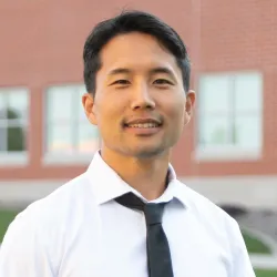 Head-and-shoulders photo of young man in white collared shirt and black tie with brick building and windows in the background