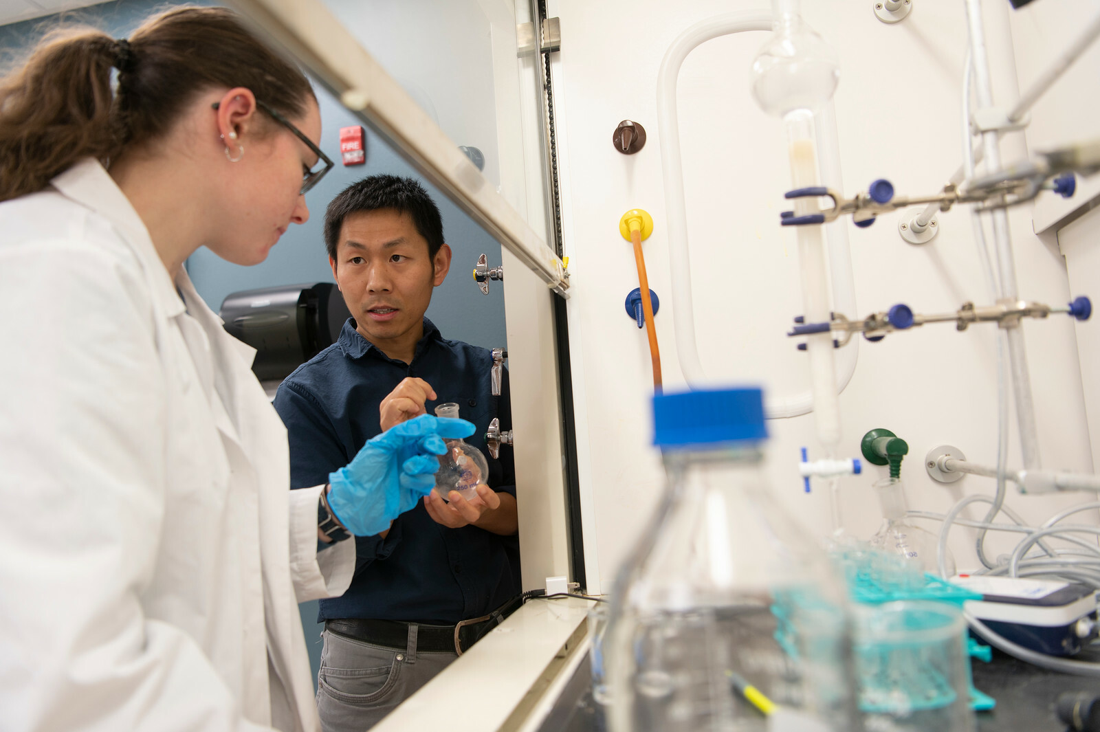 Female student in lab coat and goggles works with Dr. Zhu in the Chemistry lab
