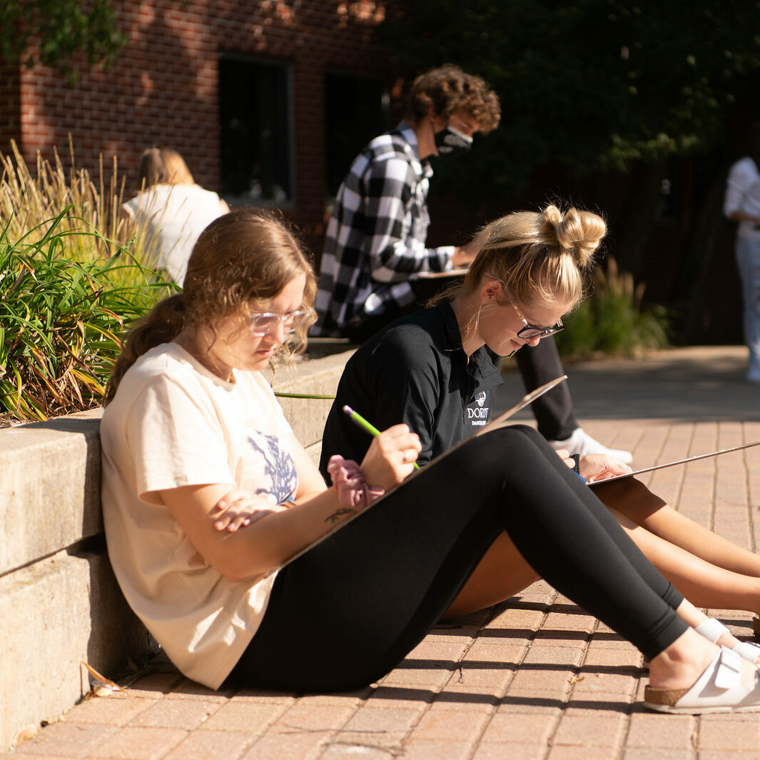 Two female students sit outside and draw next to each other
