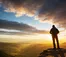 A person with a backpack stands on a cliff edge at sunrise, overlooking a scenic valley with mountains under a partly cloudy sky.