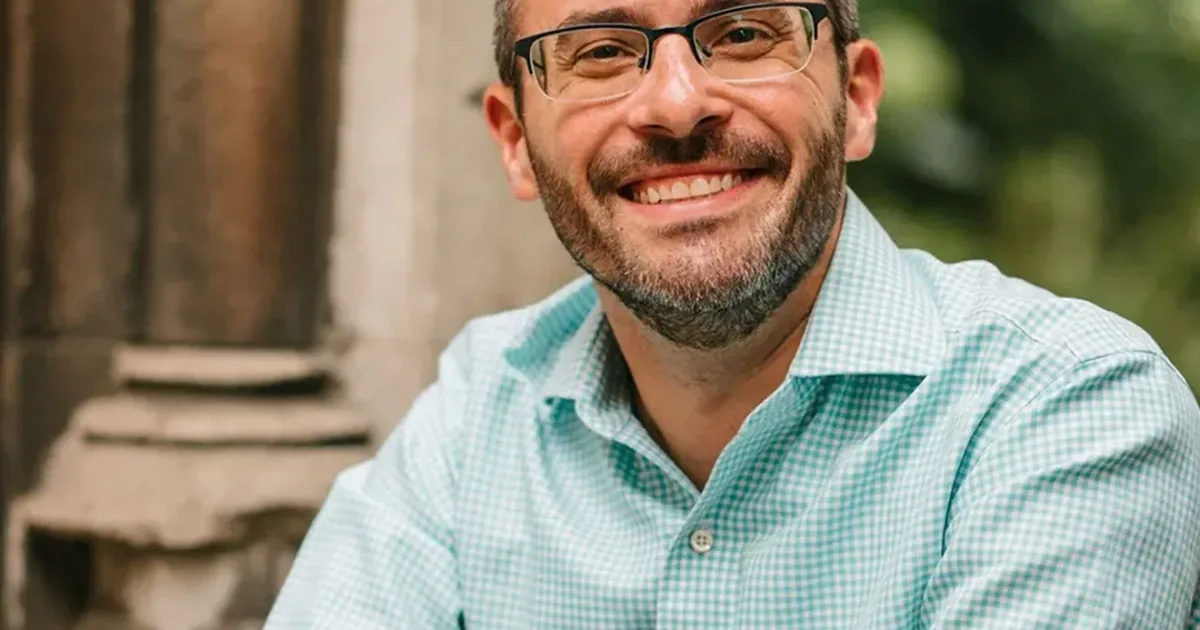 Man in collared mint-blue shirt and glasses smiles for the photo