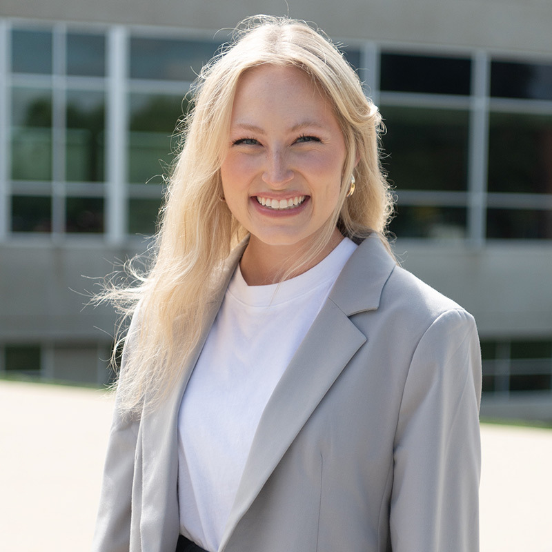 Blond-haired young woman in sport coat smiling in front of building