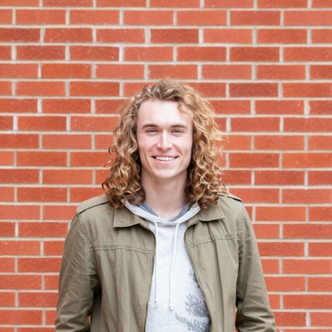 A picture of a man with long curly hair standing in front of a brick wall.